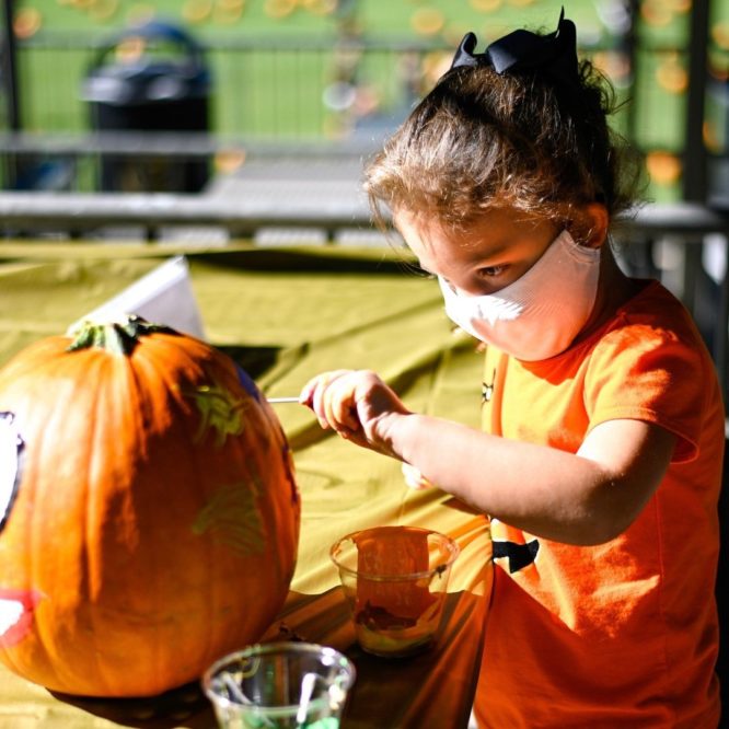 A little girl wearing orange is painting an orange pumpkin