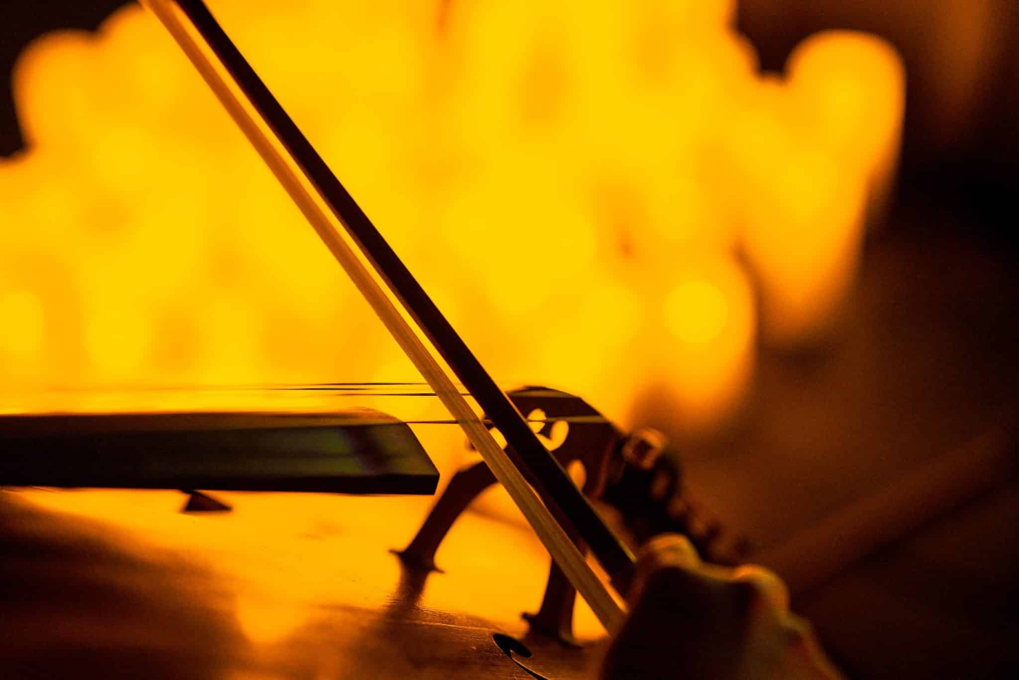 A close-up of a bow being used to play a string instrument with the blurry image of candles in the background.